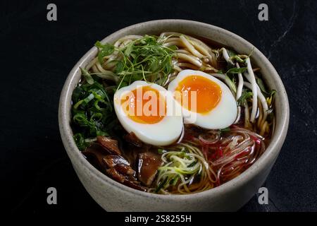 Eine Schüssel Ramen mit perfekt gekochten, weich gekochten Eiern, leuchtend grünen Zwiebeln, frischem Mikrogrün, Sojasprossen, Nudeln, und eine reiche, dunkle Brühe auf einem bl Stockfoto