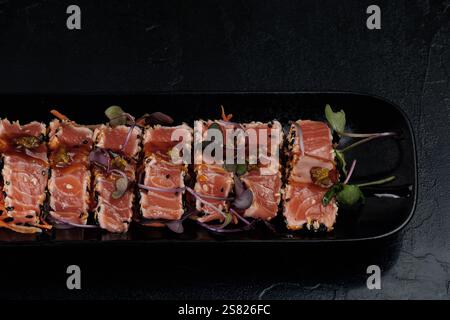 Dünne Thunfischscheiben auf einem schwarzen Teller, garniert mit Mikrogrün und Sesamsamen, verfeinert mit einer aromatischen Sauce. Die satte rote Farbe des Stockfoto