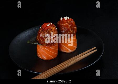 Zwei Stücke Lachs-Sushi umwickelt um scharfe Toppings, garniert mit Fischrogen und knusprigen Perlen, elegant präsentiert auf einem schwarzen Teller mit Chopstik Stockfoto