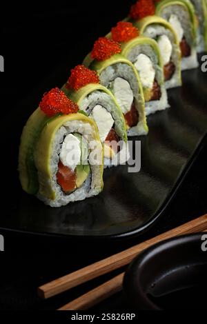 Ein wunderschön präsentierter Teller mit Sushi-Brötchen, gekrönt mit leuchtendem orangenem Fischrogen. Die Brötchen sind mit Avocado überzogen, gefüllt mit Frischkäse, Lachs, Stockfoto