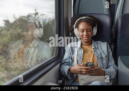 Junge afroamerikanische Frau, die Europa mit dem Zug reist und Musik über Kopfhörer hört, während sie entspannt am Fenster sitzt. Stockfoto