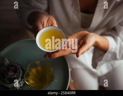 Hände halten Porzellan-Teetasse gefüllt mit goldenem Tee, der Wärme und Ruhe ausstrahlt. Elegante Einrichtung mit Teekanne und Glaskrug für eine intime Atmosphäre Stockfoto