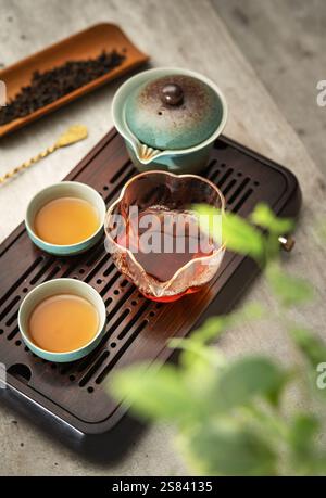 Stillleben Foto von Keramik Gaiwan, klares Glas Krug, Teetassen mit frisch gebrühtem dunklem Oolong Tee auf Tablett im Zeremoniesaal. Sanftes Licht, das Ruhe hervorruft Stockfoto