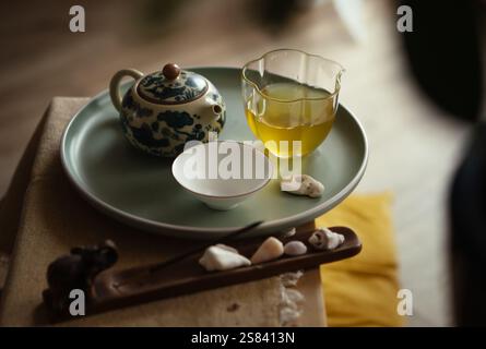 Stillleben Foto von gemusterter Teekanne aus Ton, klarem Glaskrug, Teetasse mit frisch gebrühtem Oolong-Tee auf Tablett im Zeremoniezimmer. Weiches Licht, Holztee Stockfoto