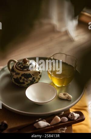 Stillleben Foto von gemusterter Teekanne aus Ton, klarem Glaskrug, Teetasse mit frisch gebrühtem Oolong-Tee auf Tablett im Zeremoniezimmer. Weiches Licht, Holztee Stockfoto