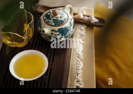 Stillleben Foto von gemusterter Teekanne aus Ton, klarem Glaskrug, Teetasse mit frisch gebrühtem grünem Oolong-Tee. Sanftes Licht, Teetablett aus Holz sorgt für beruhigende Momente Stockfoto