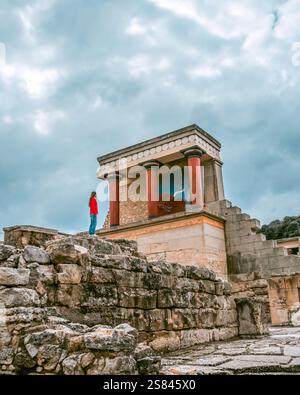 Die antiken Ruinen des Knossos Palace auf Kreta sind mit verwitterten Steinmauern, roten Säulen, einem Stierfresko und einer Person in einem roten Hemd unter bewölktem Himmel ausgestattet Stockfoto