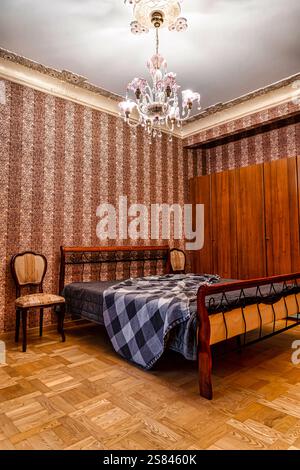 Ein Schlafzimmer im Vintage-Stil mit einem Bett mit Metallrahmen, gemusterten Tapeten, einem Holzschrank, einem Polsterstuhl und einem Blumenlüster. Stockfoto