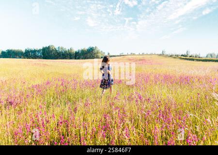 Eine Frau steht in einem lebendigen Feld aus hohem Gras und blühenden rosa Wildblumen unter einem hellblauen Himmel mit verstreuten Wolken, umgeben von fernen Bäumen Stockfoto