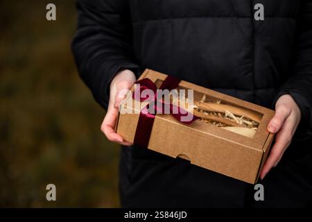 Eine Person in einer dunklen Jacke hält eine kleine Geschenkbox aus Pappe, die mit einem roten Band gebunden ist und zerfetztes Verpackungsmaterial und Holzgegenstände wie einen Löffel enthält. Stockfoto