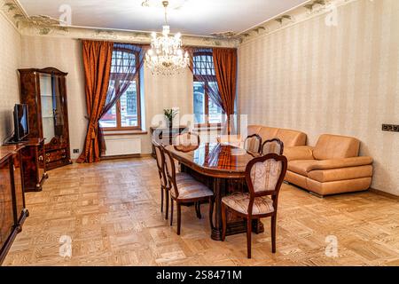 Gut eingerichtetes Wohn- und Esszimmer mit Holztisch, beigefarbenem Ledersofa, Kronleuchter, Parkettboden und gemusterten Vorhängen. Stockfoto