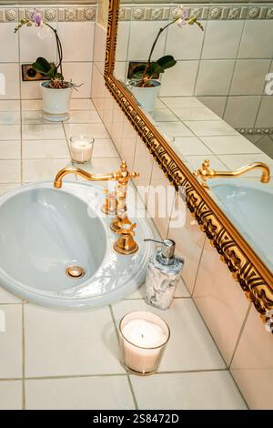 Weißes Keramikwaschbecken mit verziertem Goldhahn, beleuchteten Kerzen und einer Orchidee mit violetten Blüten auf einer gefliesten Arbeitsplatte in der Nähe eines großen goldgerahmten Spiegels Stockfoto