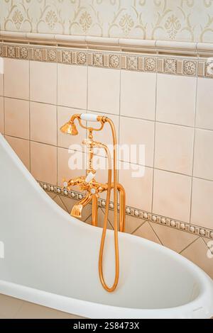 Eine weiße freistehende Badewanne mit verzierten goldenen Wasserhahn und abnehmbarer Dusche, beige und cremefarbene Fliesen mit komplizierten Mustern und Rändern. Stockfoto