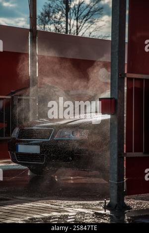 Ein schwarzes Auto wird an einer Selbstbedienungsstation gewaschen, mit Wassernebel in der Luft, roten Wänden, Metallpfählen, blattlosen Bäumen und einem teilweise bewölkten Himmel. Stockfoto