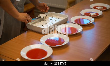 Eine Person serviert ein cremiges Dessert mit dunklen Beeren aus einem weißen Gericht auf einem Holztisch, umgeben von Tellern mit roter Sauce in einer ungezwungenen Inneneinrichtung Stockfoto