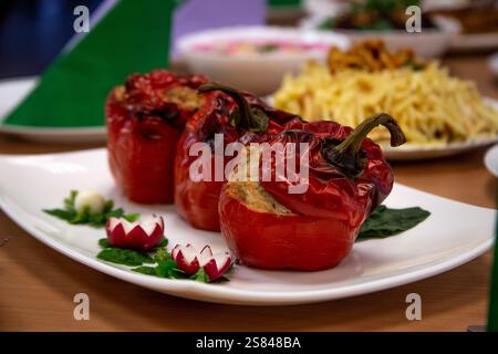 Drei gebratene rote Paprika gefüllt mit herzhafter Füllung, garniert mit frischen Kräutern und geschnitzten Radieschen, mit verschwommenen Gerichten im Hintergrund. Stockfoto