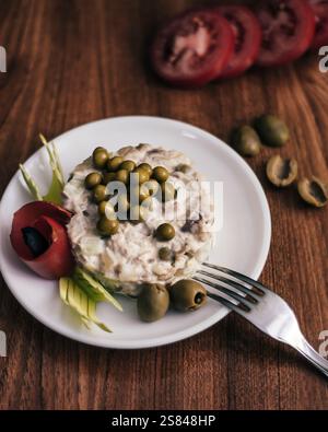 Ein runder cremiger Salat mit grünen Erbsen, garniert mit einer Tomatenrose, schwarzer Olive, Sellerieblättern und ganzen Oliven auf einer Holzoberfläche. Stockfoto