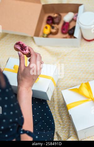 Eine Person hält einen rosafarbenen Milchteig mit roten Streuseln auf gelbem Stoff. In der Nähe gibt es buntes Gebäck, eine weiße Tasse und Geschenkboxen mit Yello Stockfoto