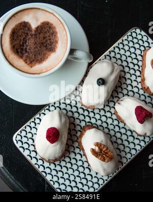 Eine Tasse Kaffee mit herzförmigem Kakaoschaum befindet sich neben einem gemusterten Teller mit sechs Backwaren, die jeweils mit Glasur und einzigartigen Garnituren belegt sind. Stockfoto