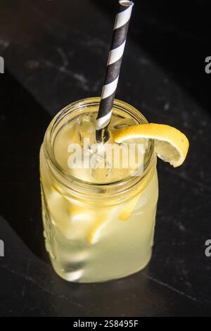 Ein mit hellgelber limonade gefülltes maurerglas mit Eiswürfeln, Zitronenscheiben und gestreiftem Stroh vor dunklem Hintergrund. Stockfoto
