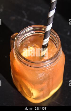 In einem maurerglas befindet sich ein goldenes Orangengetränk mit Eiswürfeln, einer Zitrusscheibe und einem schwarz-weiß gestreiften Strohhalm vor dunklem Hintergrund. Stockfoto