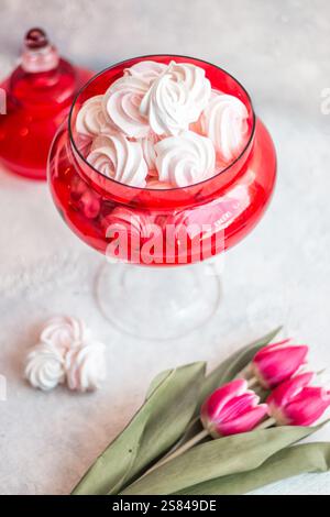 Eine rote Glasschale gefüllt mit weißen Baisons sitzt auf einer hellen Oberfläche, begleitet von rosa Tulpen und einem passenden roten Glasbehälter im Hintergrund. Stockfoto