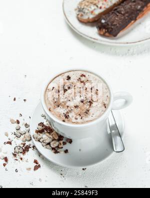 Eine weiße Kaffeetasse auf einer Untertasse mit einem silbernen Löffel, umgeben von Nuss- oder Schokoladenstücken. Zwei Eclairs mit Streuseln und Schokolade sitzen im Hintergras Stockfoto