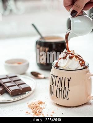 Beigefarbener Becher mit der Aufschrift TOTALLY TN CHOC, gekrönt mit Schlagsahne und Karamell, umgeben von Schokoladentafeln, Nüssen und einem Löffel auf einer hellen Oberfläche. Stockfoto