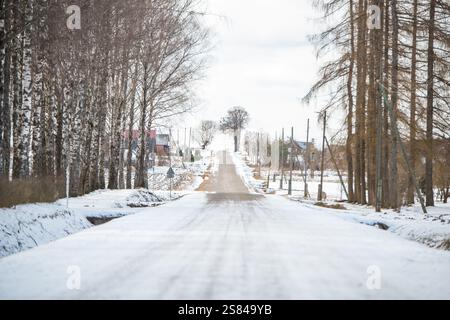 Eine winterliche Landstraße in Lettland mit Schnee, blattlosen Birken- und Nadelbäumen, einem roten Schild mit der Aufschrift „Veikals“, kargen Häusern und einem bedeckten Himmel. Stockfoto