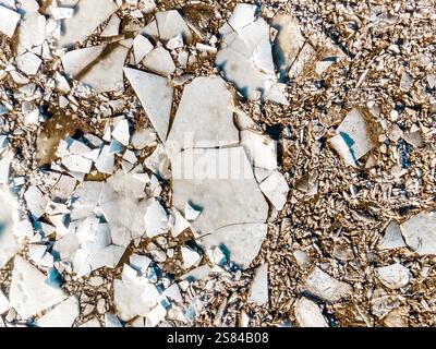 Zerrissene und gebrochene Eisschilde unterschiedlicher Größe, die über eine strukturierte Oberfläche mit kleinen kiesartigen Fragmenten und Erdtönen im Hintergrund verstreut sind Stockfoto