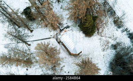 Aus der Vogelperspektive auf eine schneebedeckte Landschaft mit einer kleinen Holzhütte mit weißem Dach, kargen Bäumen und einem schmalen Holzweg. Stockfoto