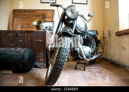 Ein Vintage-Motorrad mit sichtbarem Rost auf einem gefliesten Boden, umgeben von einer Holzkiste, einem Grammophon-Horn und gerahmten historischen Gegenständen. Stockfoto