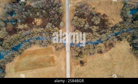 Aus der Vogelperspektive eine schmale Brücke über einen sich windenden Fluss, umgeben von dichten Bäumen und umgeben von trockenen Feldern und bewaldeten Gebieten. Stockfoto