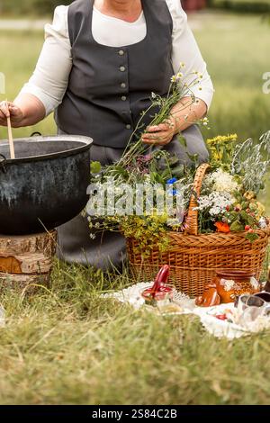 Eine Person rührt einen großen schwarzen Topf auf einem Holzstumpf auf einem grasbewachsenen Feld, mit einem Korb mit Wildblumen und einem Picknick-Set in der Nähe. Stockfoto
