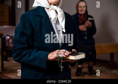 Eine ältere Frau in weißem Kopftuch und dunklem Mantel hält drinnen einen grünen Rosenkranz und ein Buch, während eine andere Frau auf einer Holzbank im Hinterhof sitzt Stockfoto