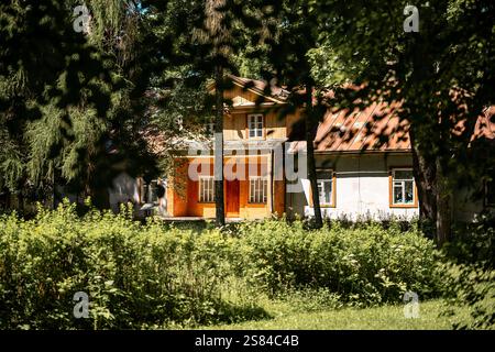 Ein rustikales Haus mit hölzernen und verputzten Wänden, einer orangefarbenen Eingangstür und weiß gerahmten Fenstern, umgeben von hohen Bäumen und üppiger Vegetation. Stockfoto