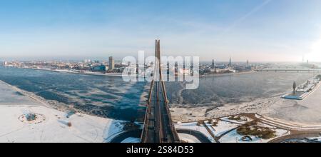 Panoramablick auf Riga, Lettland, mit der Vansu-Brücke, teilweise gefrorenem Daugava-Fluss, Türmen der Altstadt, modernen Gebäuden und schneebedeckten Gebäuden Stockfoto