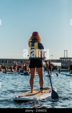 Eine Person paddelt auf ruhigem Wasser, trägt eine gelbe und blaue Schwimmweste und eine rote Kappe, andere im Hintergrund und eine Industriestruktur Stockfoto