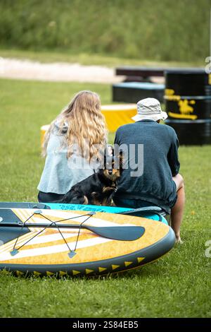 Ein Paar sitzt auf einem grasbewachsenen Feld mit einem kleinen Hund und einem farbenfrohen Paddleboard in der Nähe. Schwarze und gelbe Fässer und ein sandiger Pfad sind im Rücken sichtbar Stockfoto