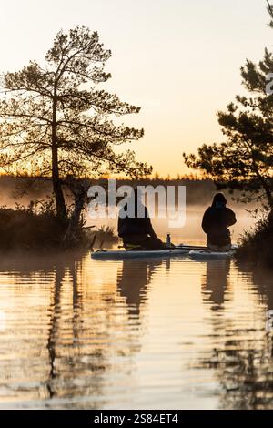 Zwei Personen sitzen auf Paddleboards in ruhigem Wasser mit aufsteigendem Nebel, eingerahmt von kargen Bäumen und goldenem Licht, das auf der Wasseroberfläche reflektiert. Stockfoto