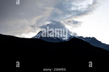 Der Berg Kasbek in Georgien erhebt sich mit schneebedeckten Gipfeln, die teilweise von Wolken bedeckt sind. Dunkle Hügel und ein dunkler Himmel verstärken die Berghöhe. Stockfoto