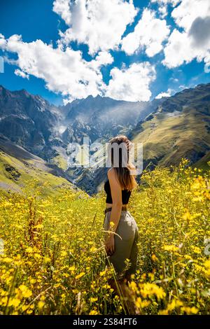 Eine Frau steht in einem Feld aus gelben Wildblumen, mit Blick auf eine zerklüftete Bergkette unter einem blauen Himmel mit verstreuten Wolken in Georgiens natürlicher Landschaft. Stockfoto