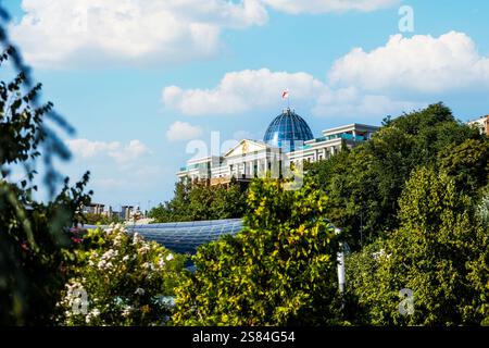 Der Präsidentenpalast in Tiflis, Georgien, verfügt über eine blaue Glaskuppel mit einer Flagge, eingerahmt von Bäumen, lebhaftem Laub und eine moderne Glasstruktur. Stockfoto