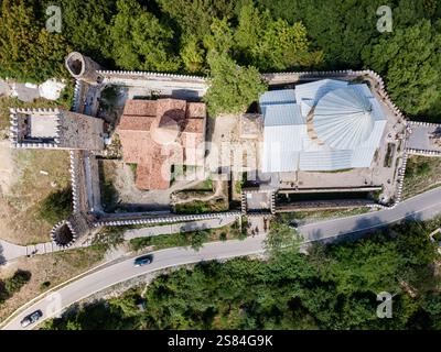 Blick aus der Vogelperspektive auf eine georgianische Festung mit Steinmauern, Türmen, roten Fliesen und Metalldächern, umgeben von Wald und einer nahe gelegenen gewundenen Straße. Stockfoto