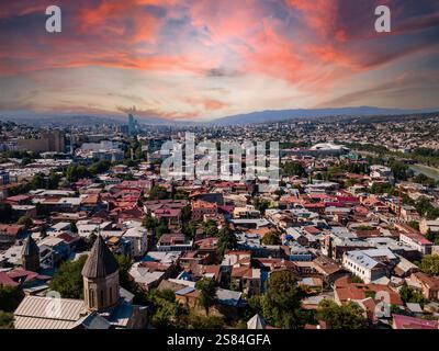 Blick auf Tiflis, Georgia, mit einer historischen Kirche, rot-weißen Dächern, dem Fluss Kura, einem modernen Wolkenkratzer und sanften Hügeln bei Sonnenuntergang. Stockfoto