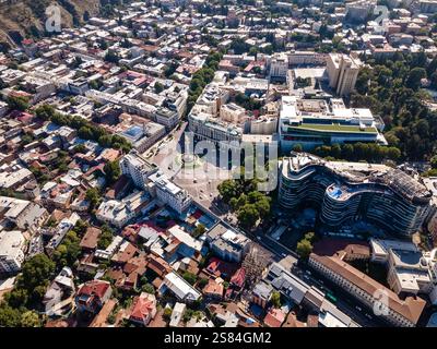 Blick aus der vogelperspektive auf einen runden platz mit einem zentralen Denkmal in Tiflis, Georgien, umgeben von modernen und historischen Gebäuden, von Bäumen gesäumten Straßen und einem Hügel Stockfoto