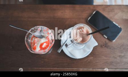 Schmelzende Eiskugel, heiße Schokolade mit Marshmallow-Belag, die neben dem Smartphone auf rustikaler Holzoberfläche liegt Stockfoto