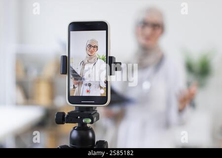 Eine muslimische Ärztin mittleren Alters nimmt medizinische Videostunden mit dem Smartphone auf. Konzentriert sich auf Gesundheitswesen, Bildung und Technologie. Zeigt Röntgenaufnahmen und Behandlungserklärungen an. Stockfoto