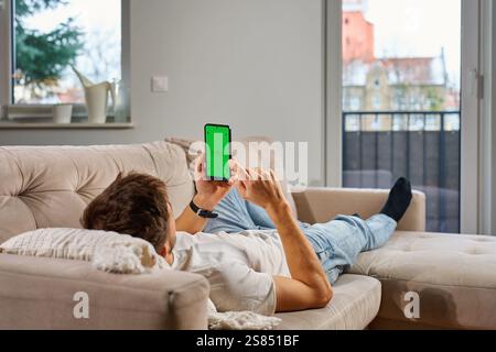 Ein Mann in lässiger Kleidung liegt auf der beigen Couch, hält ein Smartphone mit grünem Bildschirm und entspannt sich im modernen Wohnzimmer. Mann, der sich auf dem Sofa ausruht und sich anschaut Stockfoto
