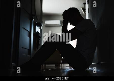 Depressiver junger Mann, der auf dem Boden in einem dunklen Zimmer sitzt. Silhouette eines jungen Mannes, der an Einsamkeit und Angst leidet und sein Gesicht mit Händen bedeckt. Perso Stockfoto